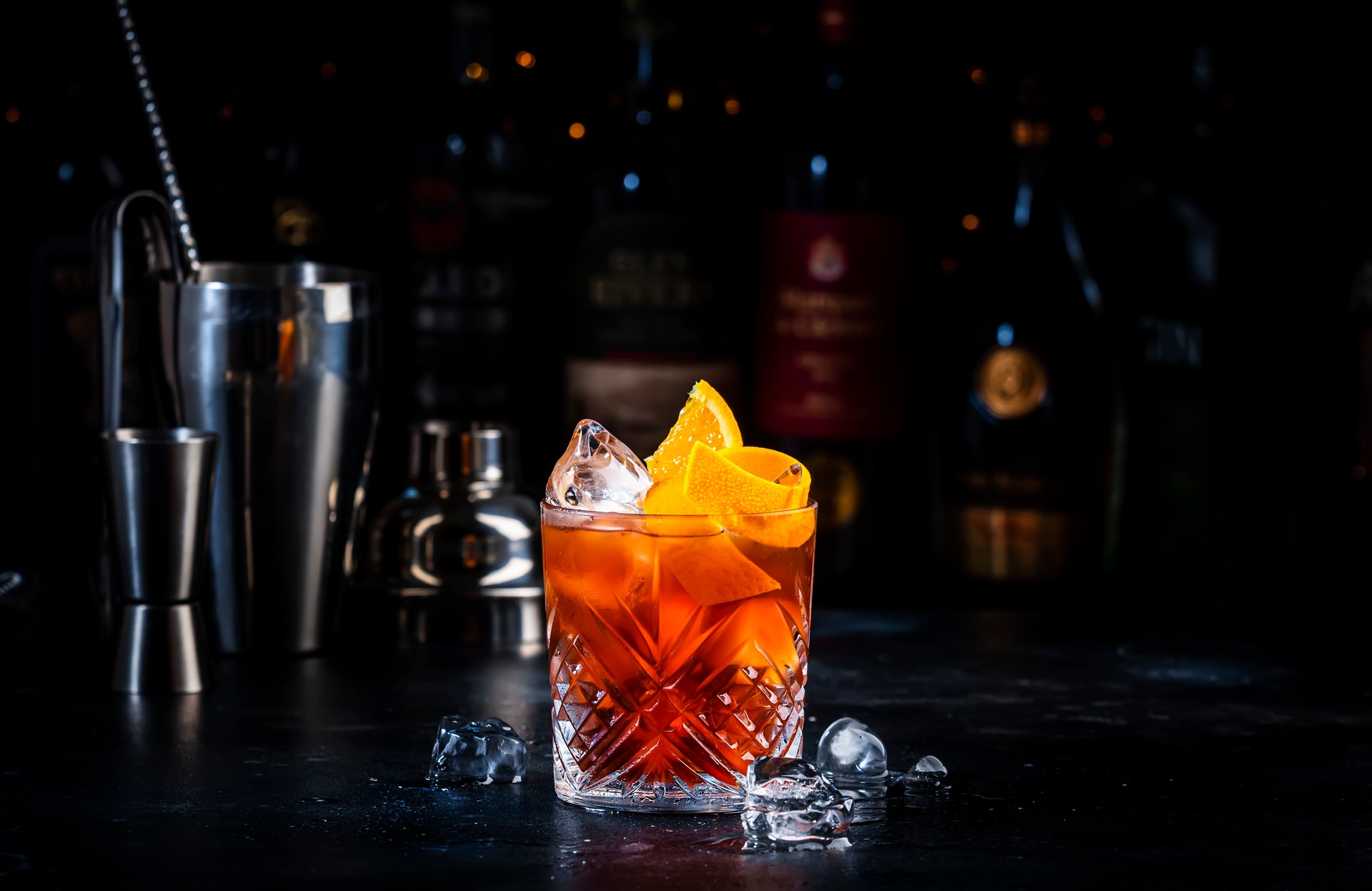 Espresso Negroni cocktail drink with dry gin, red vermouth and bitter, espresso, coffee liqueur, orange zest and ice. Black bar counter background, bar tools, bottles